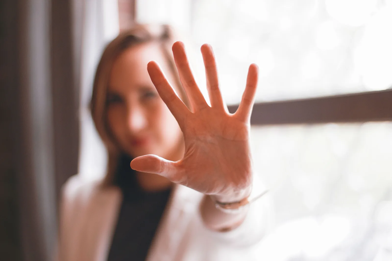 Eine Frau hält ihre geöffnete Hand mit fünf gespreizten Fingern in die Kamera. Die Hand ist scharf, ihr Gesicht und das Fenster dahinter sind unscharf.