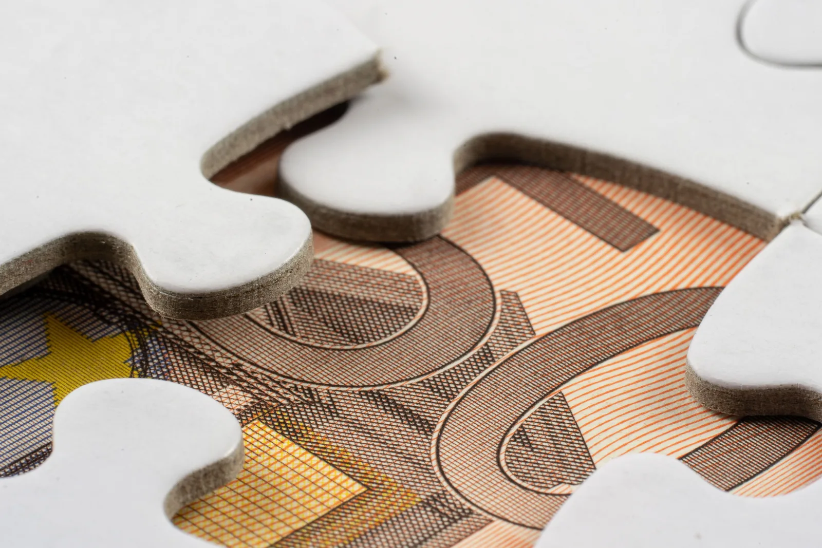 Weiße Puzzleteile liegen auf einer Euro-Banknote und verdecken sie zum Teil. Durch die Lücken sind Ziffern und das Guillochenmuster des Scheins zu sehen.